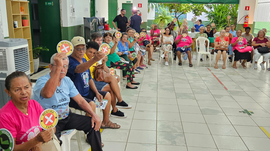 Imagem principal mostra grupo de idosos e idosas reunidos em um espaço coletivo, sentados em cad...