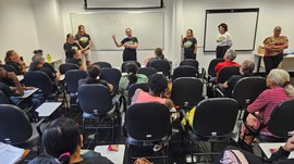 A imagem mostra uma sala de aula com adultos e idosos sentados em cadeiras escolares, atentos à ...