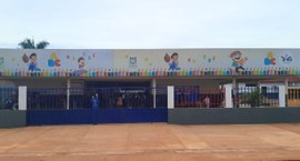 A imagem mostra a fachada de uma escola municipal cercada por grades coloridas, com portão azul ...