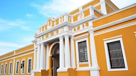 A imagem mostra a fachada de um prédio histórico pintado em amarelo com detalhes em branco. A co...