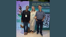 Três representantes do TRE-MT posam para foto em frente ao painel do 6º Encontro Nacional de Mem...