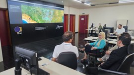 A imagem mostra uma sala do TRE-MT, com um grande painel digital exibindo um mapa de Mato Grosso...