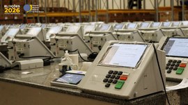 A imagem mostra diversas urnas eletrônicas modelo UE2022 organizadas em fileiras sobre bancadas,...